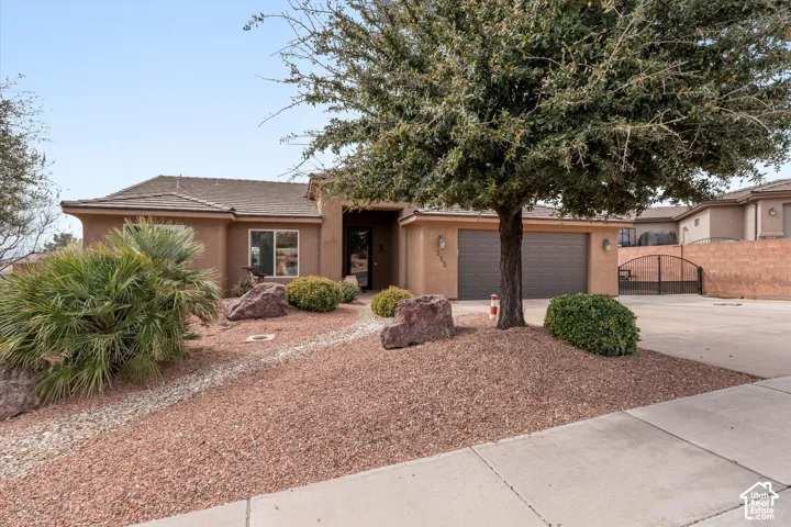 Stucco exterior featuring a dual-car garage, low-maintenance desert landscaping, mature tree, and decorative rock hardscaping