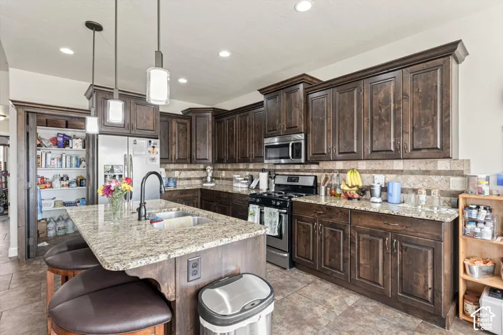 Kitchen featuring dark wood cabinetry, stone-surface countertops, stone-tile backsplash, central island with an integrated stainless steel dual-basin sink, and recessed lighting