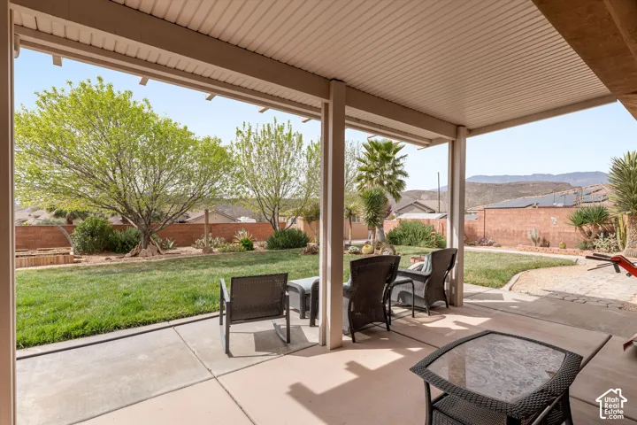 Covered concrete patio featuring a solid-surface ceiling, expansive grass lawn, masonry perimeter wall, mature trees, and mountain backdrop