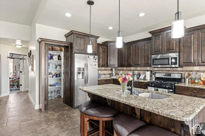 Kitchen featuring dark-stained wood cabinetry, stone countertops, stainless steel appliances, a center island with an integrated sink, and tile flooring