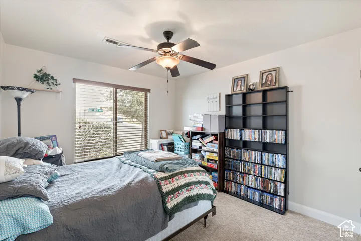 Neutral-toned bedroom featuring plush carpeting, an overhead ceiling fan with integrated lighting, and a large window fitted with horizontal blinds
