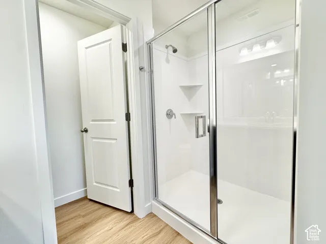 Walk-in shower featuring a white surround, integrated shelving, and a chrome-finish shower head, framed by a glass door with a chrome handle, complemented by wood-finish flooring and a white paneled door
