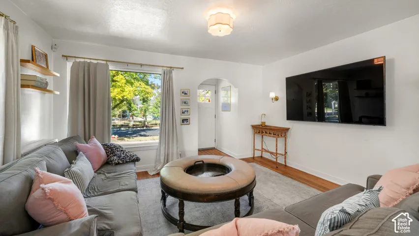 Living space featuring wood-finish flooring, a large picture window, arched entryway, wall-mounted light fixtures, and wall-mounted floating wood shelves