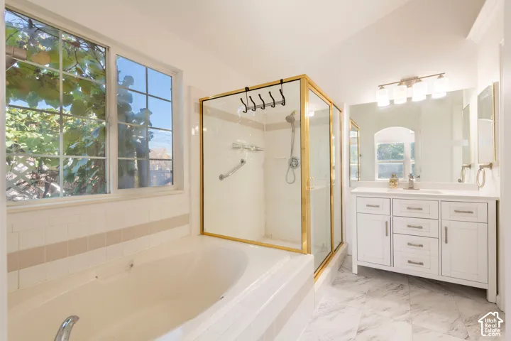 Bathroom featuring a white vanity cabinet with stone-finish countertop, a separate deep soaking tub, and a walk-in shower with gold-tone metal framing