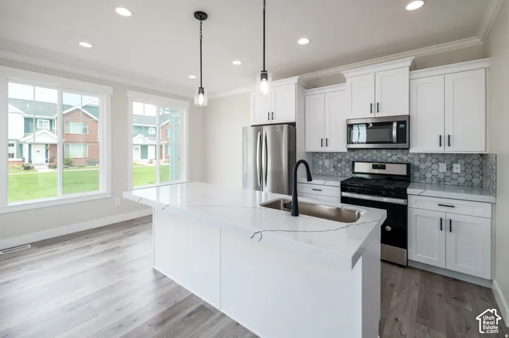 Open-concept kitchen featuring white cabinetry, stainless steel appliances, a tile backsplash, light wood-finish flooring, and a central island with an undermount sink and pendant lighting