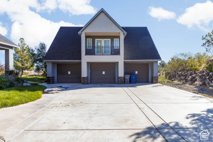 Three-car garage exterior featuring a stucco facade, stone accents, a private upper-level balcony with iron railing, and an expansive concrete driveway