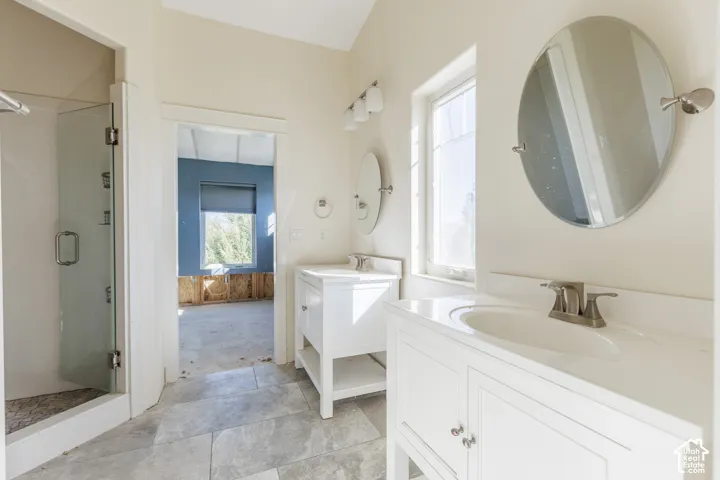 Dual white vanities with integrated sinks, glass-enclosed shower, stone-finish tile flooring, and round wall-mounted mirrors