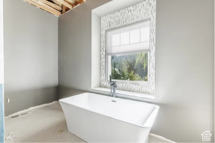 Freestanding white soaking tub featuring a deck-mounted chrome faucet, set against a window framed by vertical mosaic tile backsplash