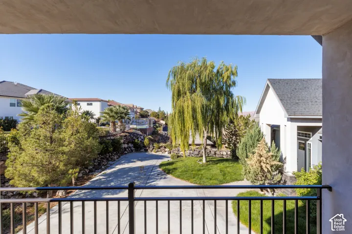 Covered balcony featuring black metal railings, overlooking a landscaped grounds area with mature willow trees, stone retaining walls, and concrete paving