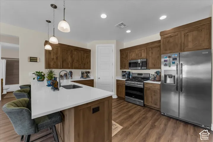 Kitchen featuring wood cabinetry, white countertops, stainless steel appliances, a center island with an undermount sink, and pendant lighting