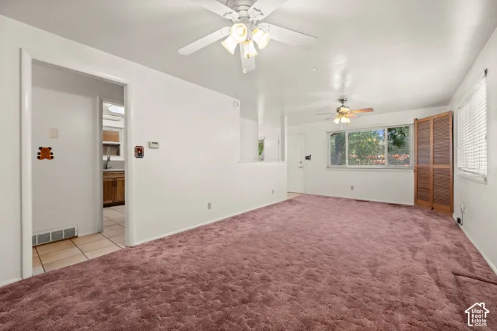Living area featuring dual ceiling fans with integrated lighting, wall-to-wall carpeting, large window with horizontal blinds, and wood-finish folding doors