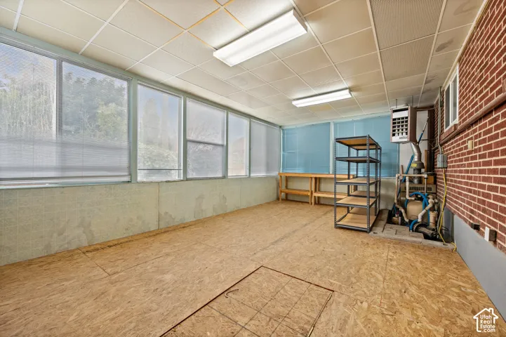 Enclosed sunroom featuring expansive windows, exposed brick walling, a drop ceiling with fluorescent lighting, and industrial-style utility access