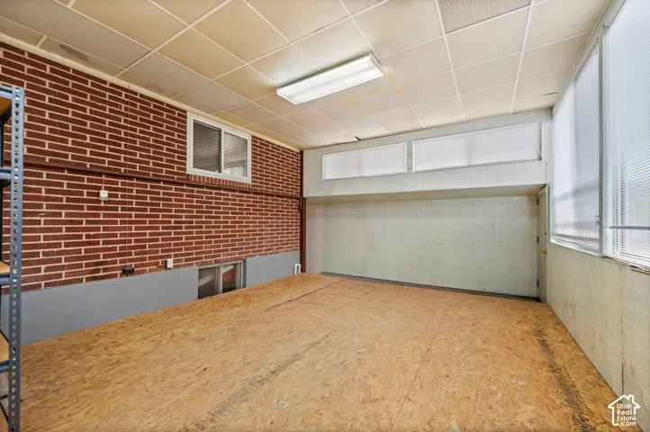 Interior space featuring a brick accent wall, dropped ceiling with integrated lighting, wood-composite flooring, and dual-pane window installations