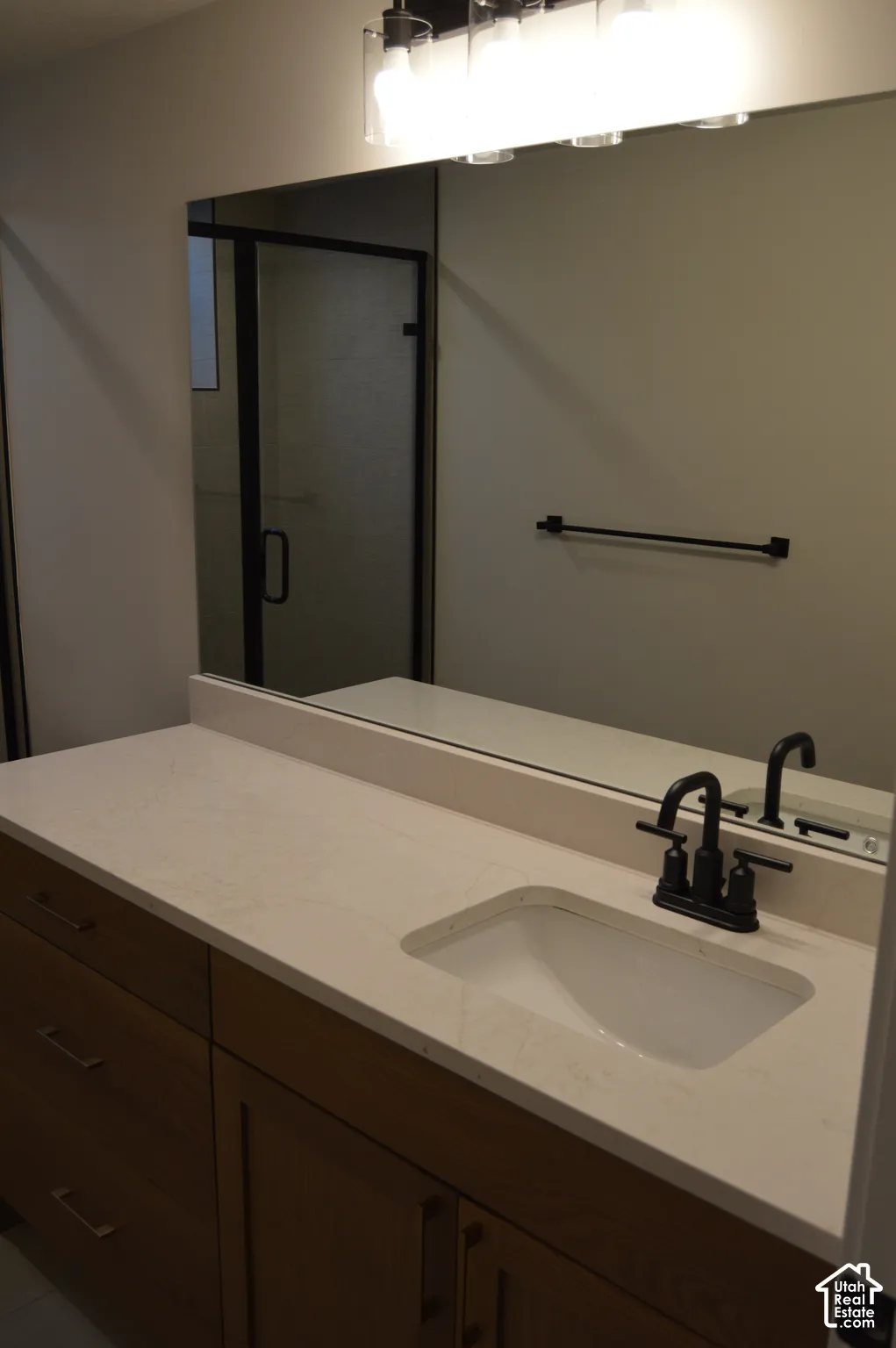 Vanity featuring wood-finish cabinetry, a neutral countertop with an integrated basin, matte black faucet hardware, and a large-format mirror with multi-bulb overhead lighting