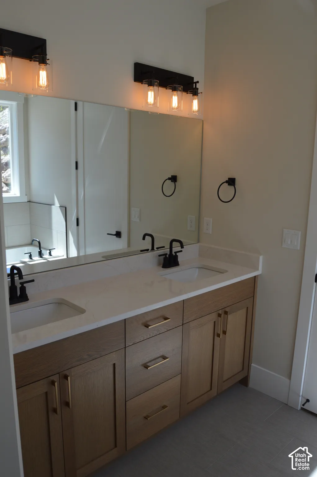 Dual-sink vanity with light-wood finish cabinetry, rectangular under-mount sinks, stone-finish countertops, matte black plumbing fixtures, and wall-mounted industrial-style light fixtures