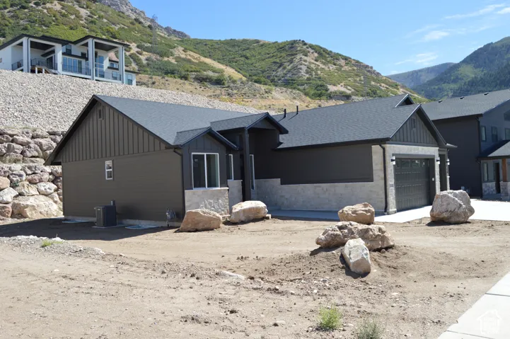 Exterior featuring dark vertical and horizontal siding, stone veneer accents, an asphalt shingle roof, and a multi-car garage, set against a mountainous backdrop