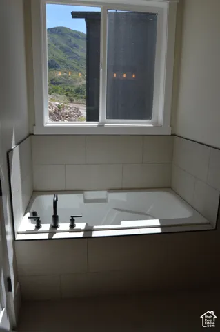 Soaking tub with a deck-mounted matte black faucet, surrounded by large-format neutral tile and a sliding window with mountain scenery