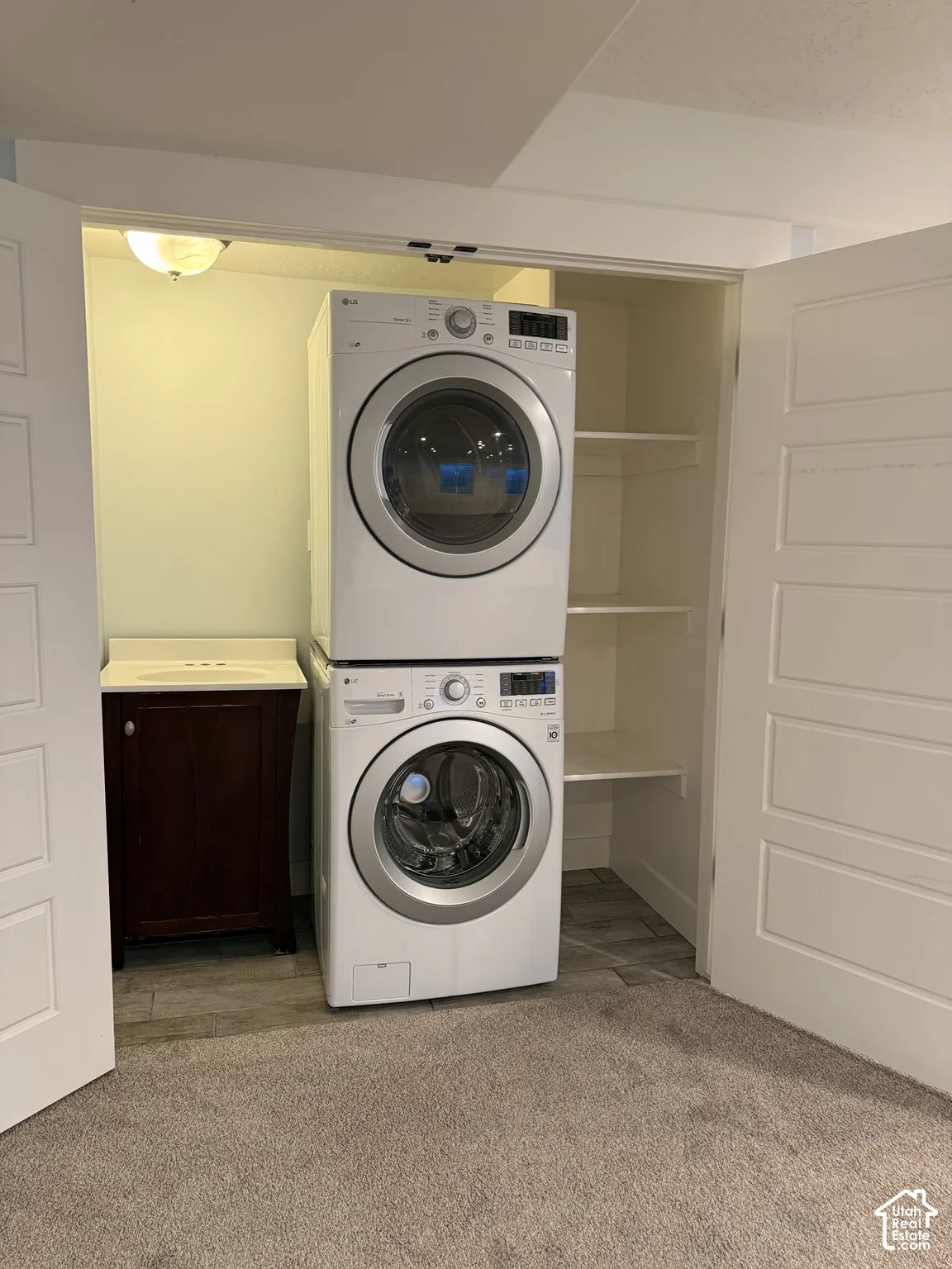 Laundry closet featuring stacked front-load appliances, a dark wood-finish vanity with an integrated sink, built-in shelving, and neutral wall-to-wall carpeting