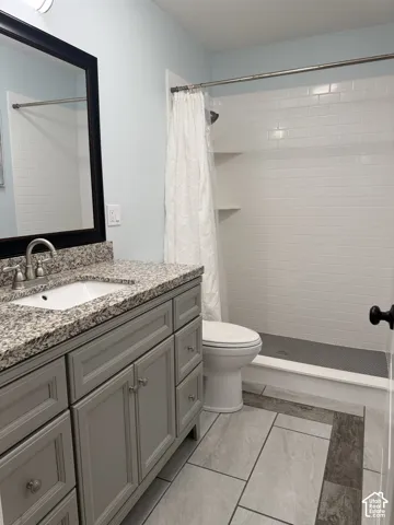 Bathroom featuring a grey shaker-style vanity with stone-patterned countertop, under-mount sink, tiled shower surround with built-in shelving, and large-format tile flooring