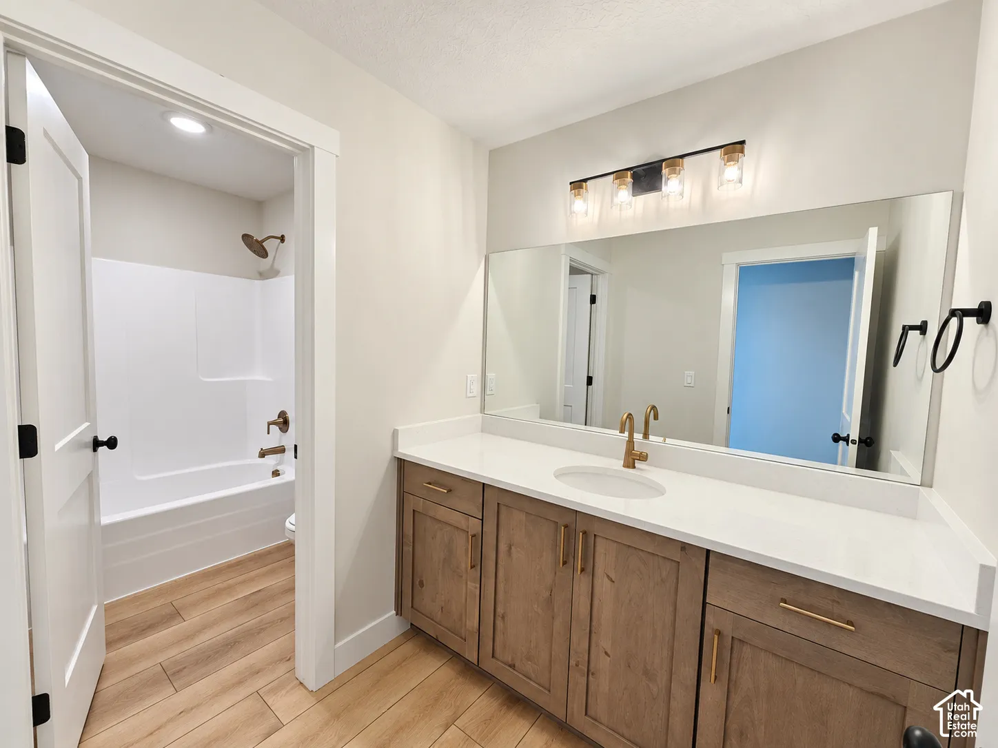 Bathroom vanity featuring wood-finish cabinetry, solid-surface countertop, integrated sink, and brass-tone hardware, paired with a shower-tub combination featuring matching brass-tone fixtures and wood-finish flooring