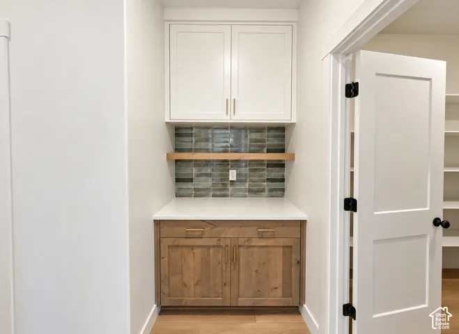 Built-in cabinetry featuring wood-finish lower cabinets, white upper cabinets, quartz countertop, floating wood shelf, and decorative tile backsplash with metallic hardware