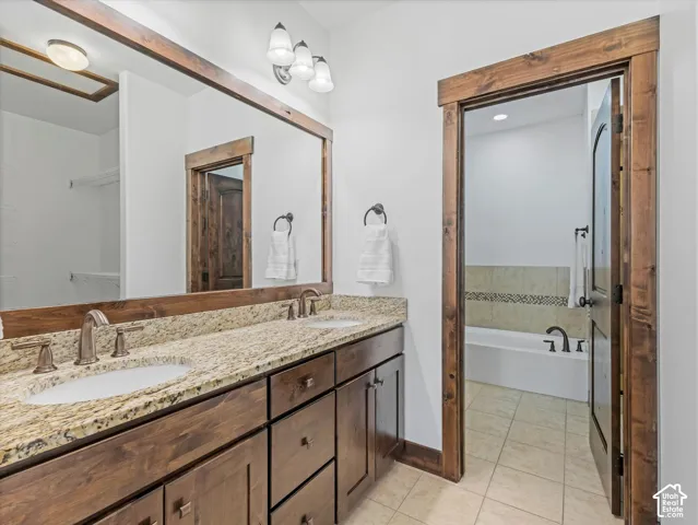 Dual-sink vanity with stone countertop, stained wood cabinetry, framed mirror, and brushed bronze fixtures, featuring wood-trimmed doorways and tiled flooring