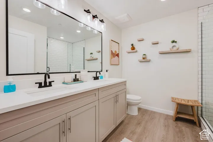 Dual-sink vanity with light-tone cabinetry, stone-surface countertop, matte black fixtures, full-width mirror, and wood-finish flooring