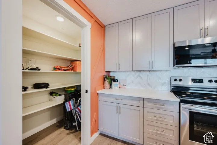 Kitchen featuring grey shaker cabinetry, white solid-surface countertops, herringbone-patterned tile backsplash, stainless steel appliances, and a walk-in pantry with wood-finish flooring