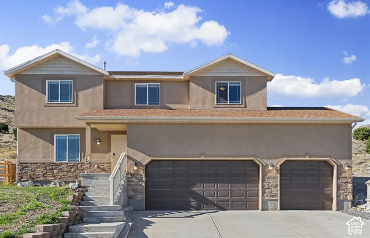 Two-story residence featuring a tan stucco and stone veneer exterior, an attached three-car garage, shingle roofing, and concrete entry stairs