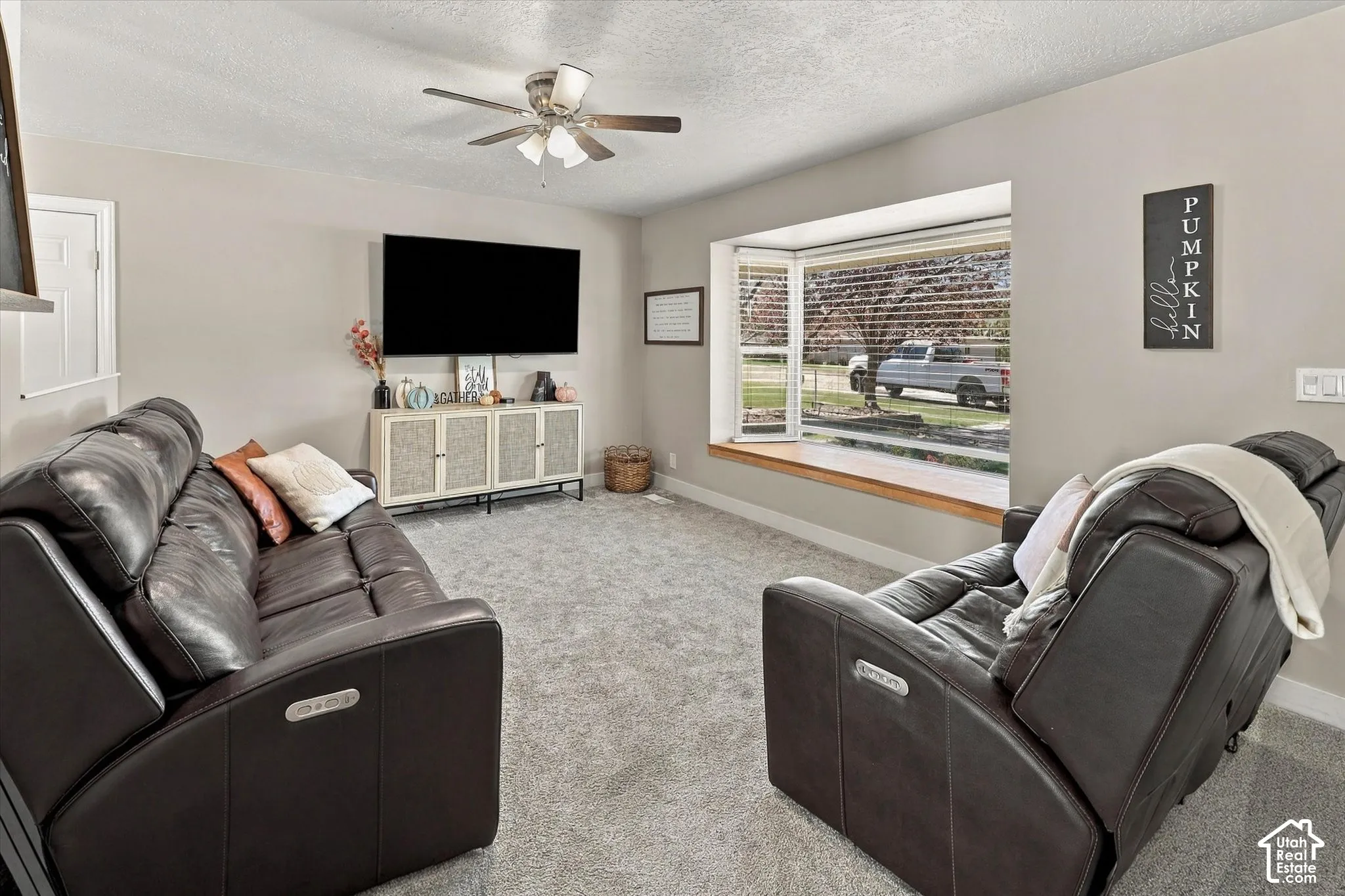 Living area featuring neutral wall tones, wall-to-wall carpeting, a multi-blade ceiling fan with integrated lighting, and a prominent bay window with a wooden sill