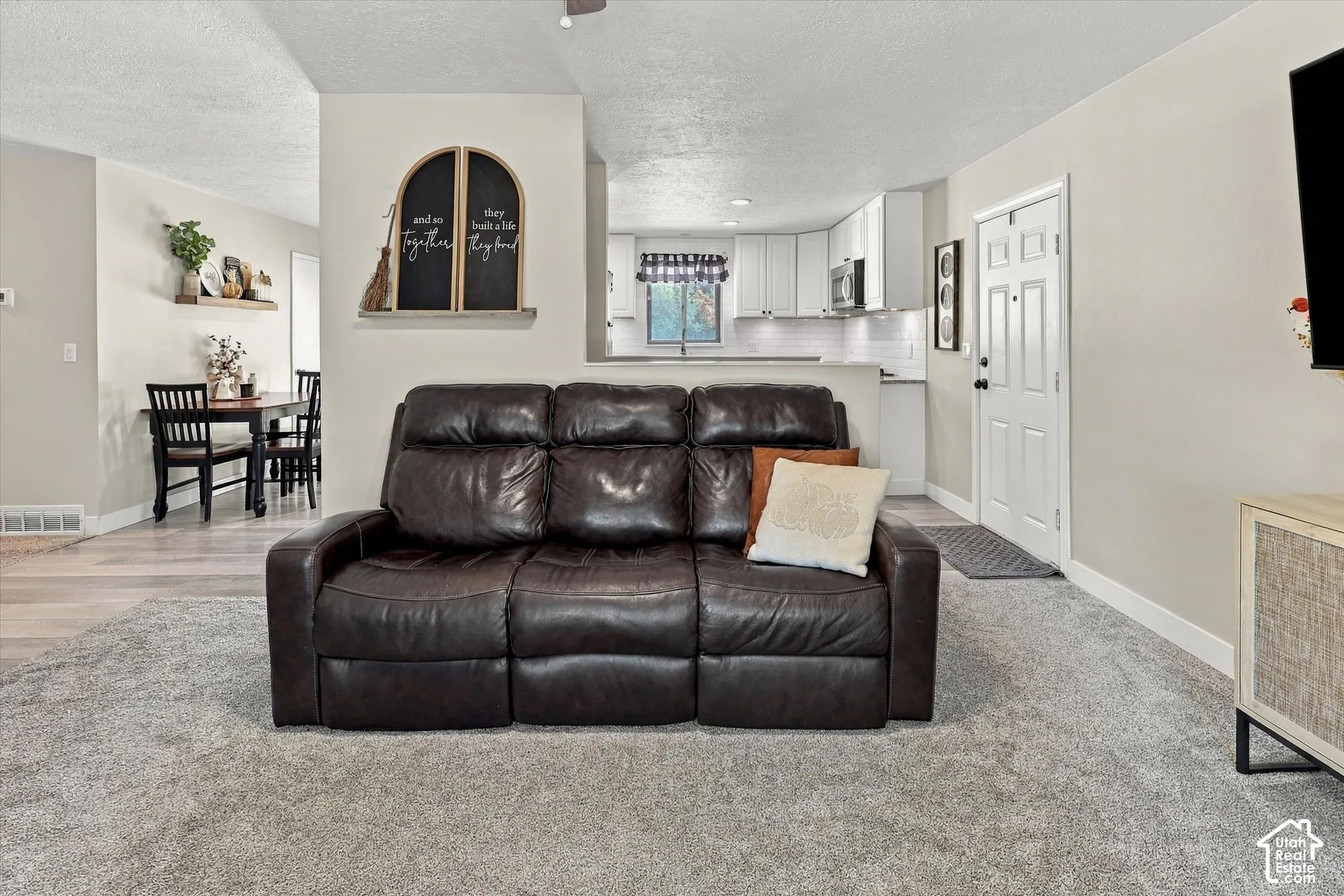 Open-concept interior featuring neutral wall tones, carpeted flooring, wood-finish flooring, white kitchen cabinetry, and a six-panel entry door