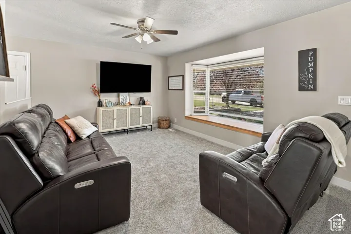 Living area featuring neutral wall tones, wall-to-wall carpeting, a multi-blade ceiling fan with integrated lighting, and a prominent bay window with a wooden sill