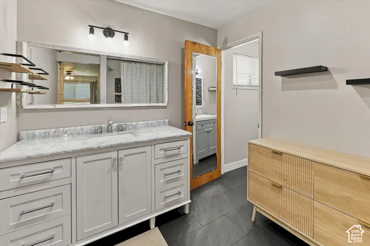 Bathroom featuring a gray vanity with stone-style countertop and integrated sink, a rectangular mirror with a white distressed wood frame, dark tile flooring, and wall-mounted shelving