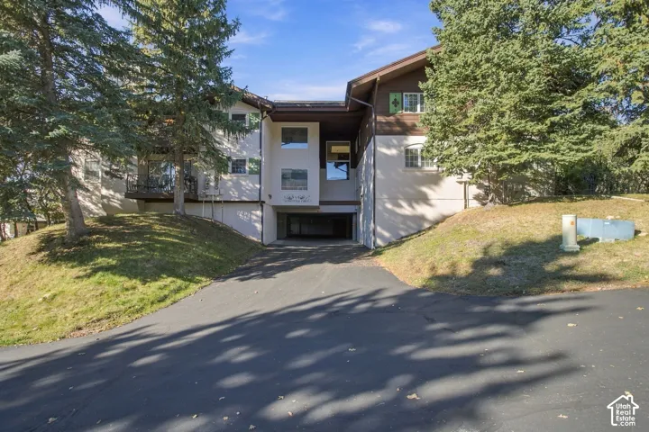 Multi-level exterior featuring stucco and wood-paneled siding, integrated covered parking, elevated balcony, and mature coniferous landscaping