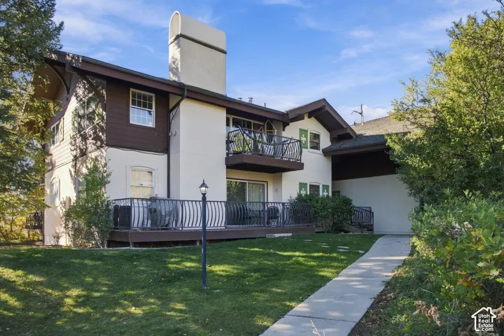 Two-story residence featuring light-colored stucco siding, dark wood trim, an elevated balcony with decorative metal railing, a ground-level deck with matching metal balustrades, and a central masonry chimney