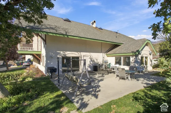 Concrete patio, upper-level balcony, shingled roof, sliding glass doors, and manicured lawn