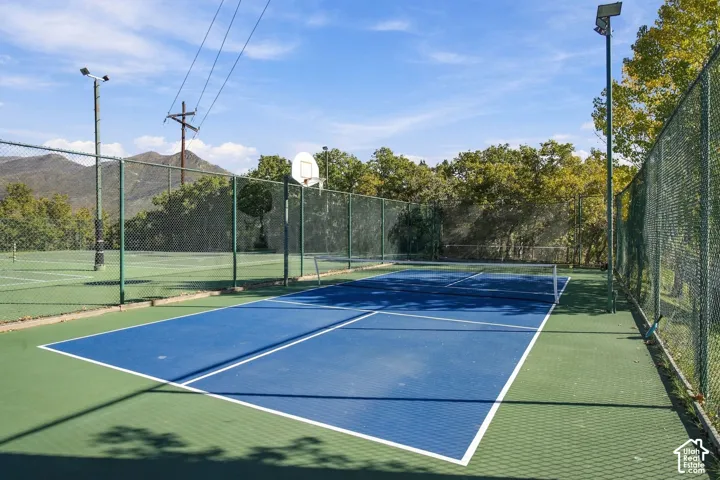 Sport court featuring a multi-use surface, basketball hoop, tennis net, perimeter chain-link fencing, and overhead lighting