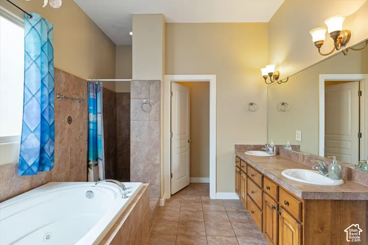 Bathroom featuring a jetted soaking tub, dual-sink vanity with tile countertop, tile flooring, and wall-mounted vanity lighting