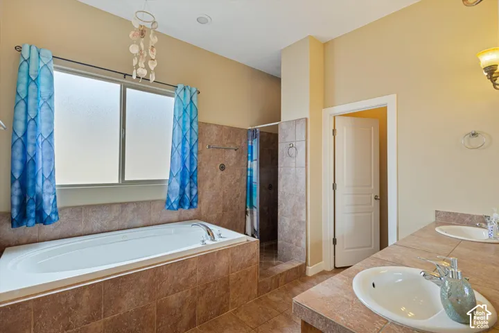 Bathroom featuring a jetted soaking tub with tile surround, dual undermount sinks, tiled countertop, and a walk-in shower