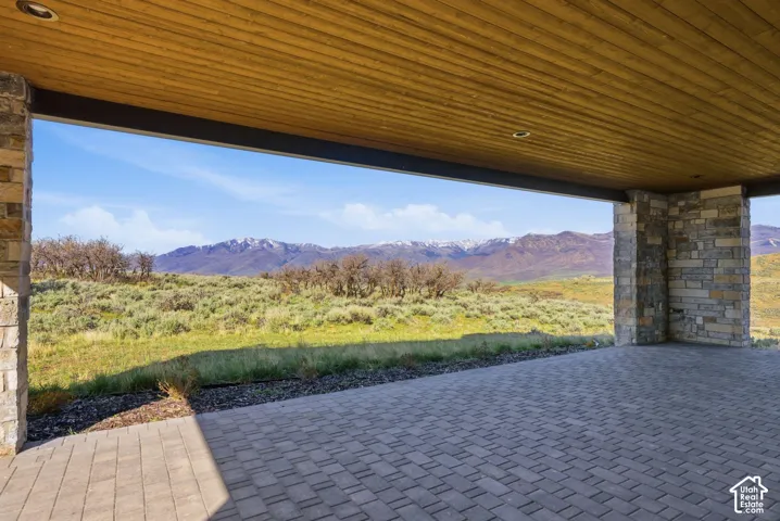Expansive covered patio featuring a paver surface, stone-clad columns, and a wood-finish ceiling with recessed lighting, all overlooking a natural landscape with distant mountains