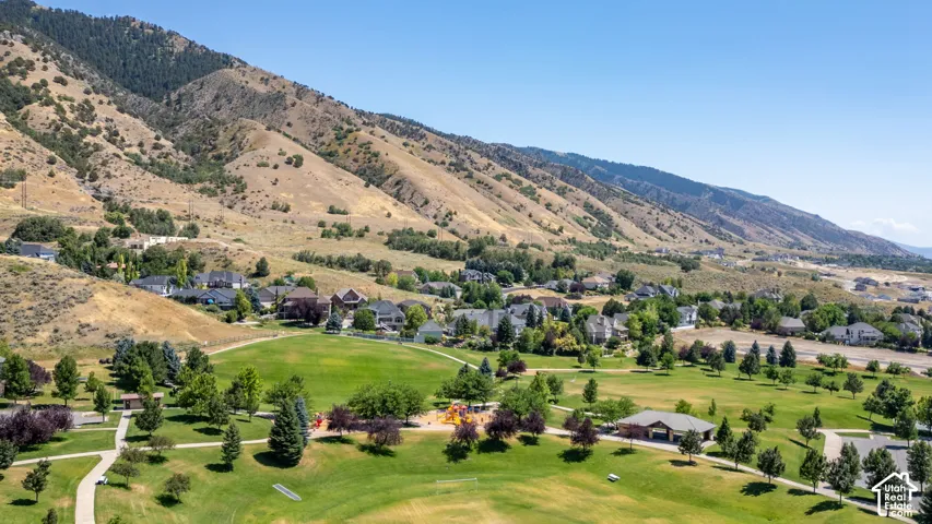 Aerial landscape featuring mountain terrain, expansive grass fields, paved pathways, mature trees, and a covered pavilion