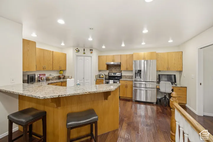 Kitchen featuring wood cabinetry, stone countertops, wood-finish flooring, stainless steel appliances, and a peninsula with breakfast bar seating