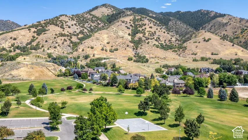 Aerial perspective of an expansive neighborhood bordered by mountainous terrain, featuring manicured green space, paved pathways, and a concrete sports court