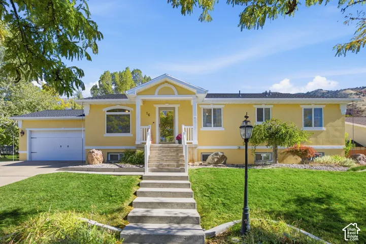 Single-story residence featuring yellow stucco siding, a gabled entryway with a white railing, an attached garage, and landscaped front grounds with stone accents