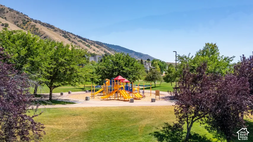 Outdoor recreational area featuring a playground structure, mature trees, manicured lawns, and a mountain backdrop