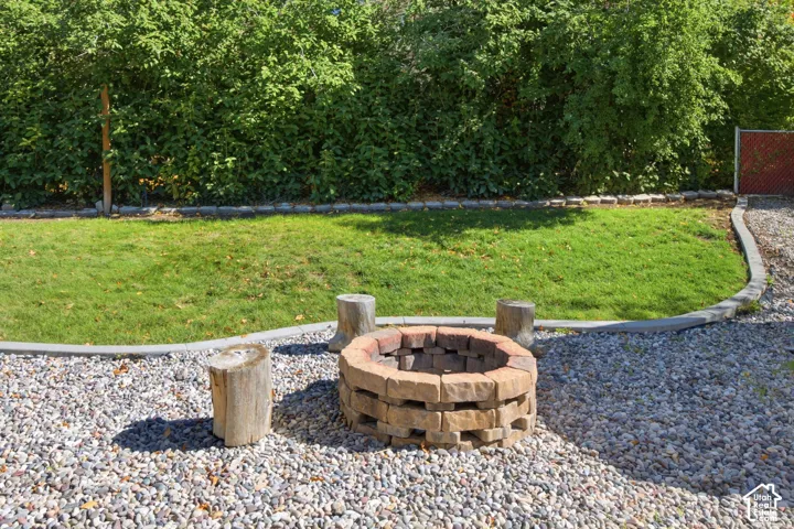 Backyard area featuring a circular stone fire pit, gravel ground cover, manicured lawn, concrete paver edging, and perimeter greenery