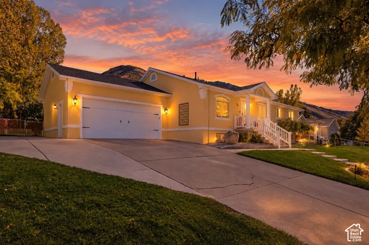 Stucco exterior with a two-car garage, concrete driveway, covered porch with white railing, and mountainous backdrop