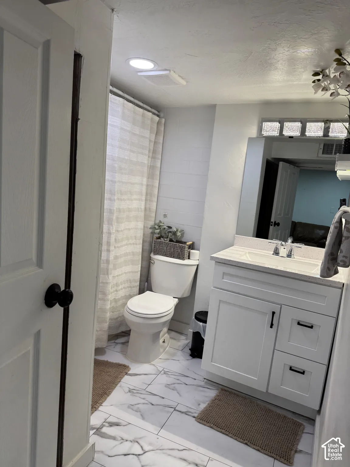Bathroom in basementbfeaturing white shaker-style cabinetry with matte black hardware, stone-finish tile flooring, a rectangular vessel sink with a polished chrome faucet, and a large wall-mounted mirror with an integrated glass block accent