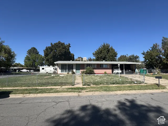 Single-story exterior featuring a brick facade, low-pitch roofline, attached carport, and chain-link perimeter fencing