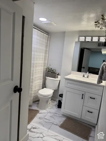 Bathroom in basementbfeaturing white shaker-style cabinetry with matte black hardware, stone-finish tile flooring, a rectangular vessel sink with a polished chrome faucet, and a large wall-mounted mirror with an integrated glass block accent